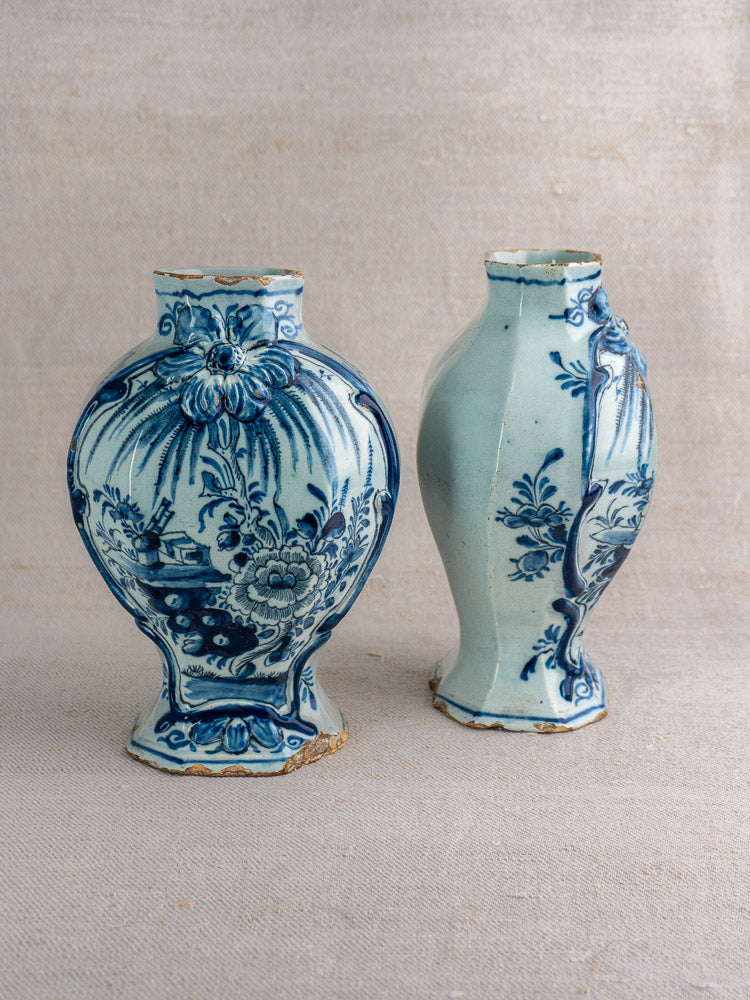 Antique Dutch Delft vase pair, blue and white with floral motifs, visible wear and chipping, on neutral fabric background.