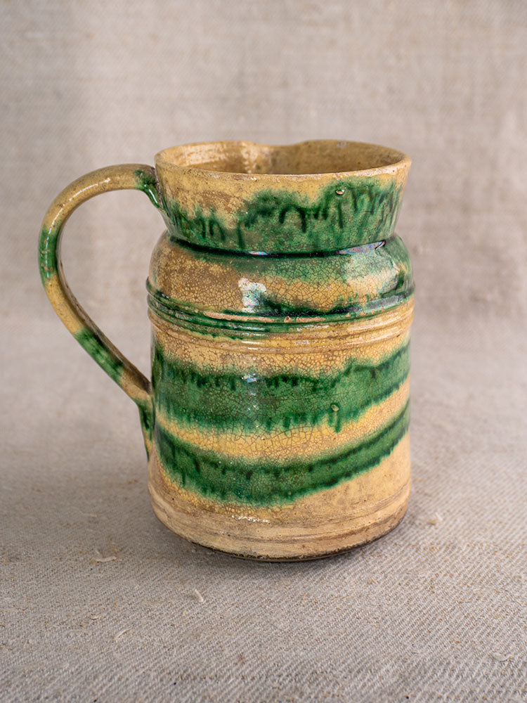 Pair of antique French Jaspé pitchers with green glaze, textured surfaces, and rustic handles on beige fabric background.
