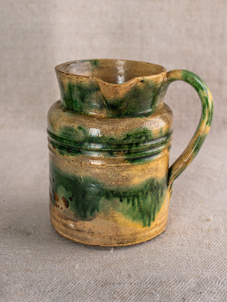 Pair of antique French jaspé Dieulefit pitchers with green glaze, brown bases, wavy rims, on neutral fabric background.