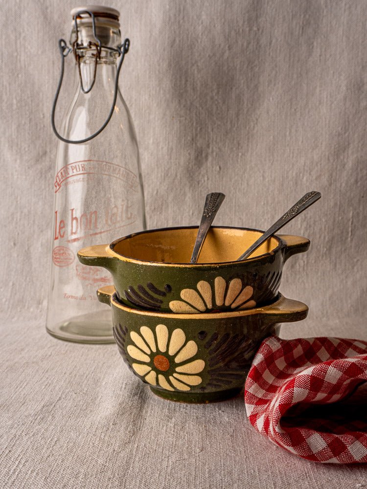Stacked green glazed Savoyard bowls with handles and spoons beside a “Le bon lait” milk bottle and red checkered cloth.