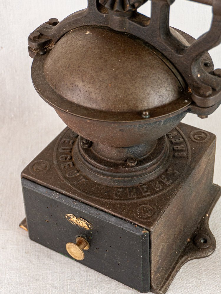 Antique French Peugeot Frères cast iron coffee mill, dome top and small drawer, on light fabric.