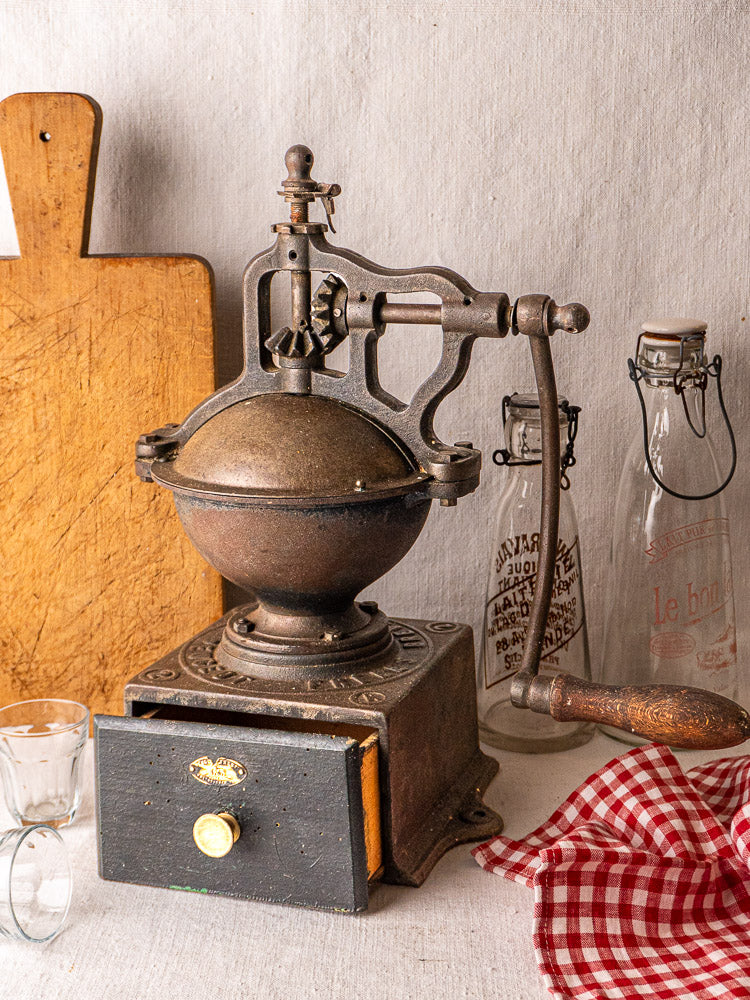 Antique French Peugeot Frères cast iron coffee mill with round top, crank handle, and drawer; light gray background.