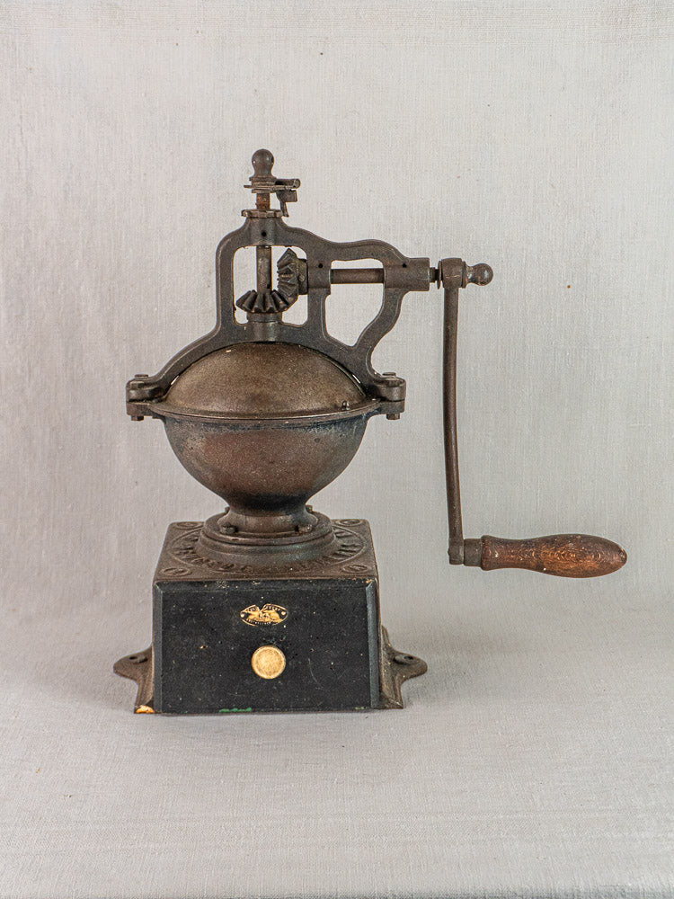 Antique French Peugeot Frères cast iron coffee mill with round top, crank handle, and drawer; light gray background.