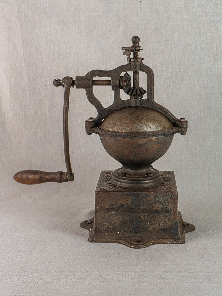 Antique French Peugeot Frères cast iron coffee mill with round bowl, crank handle, and square base on neutral fabric.