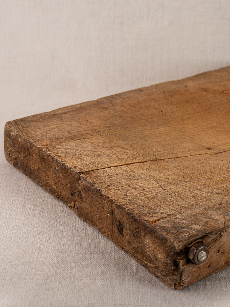 Antique French hand-carved cutting board, early 19th century, weathered wood with cracks and rusty bolt on fabric background.
