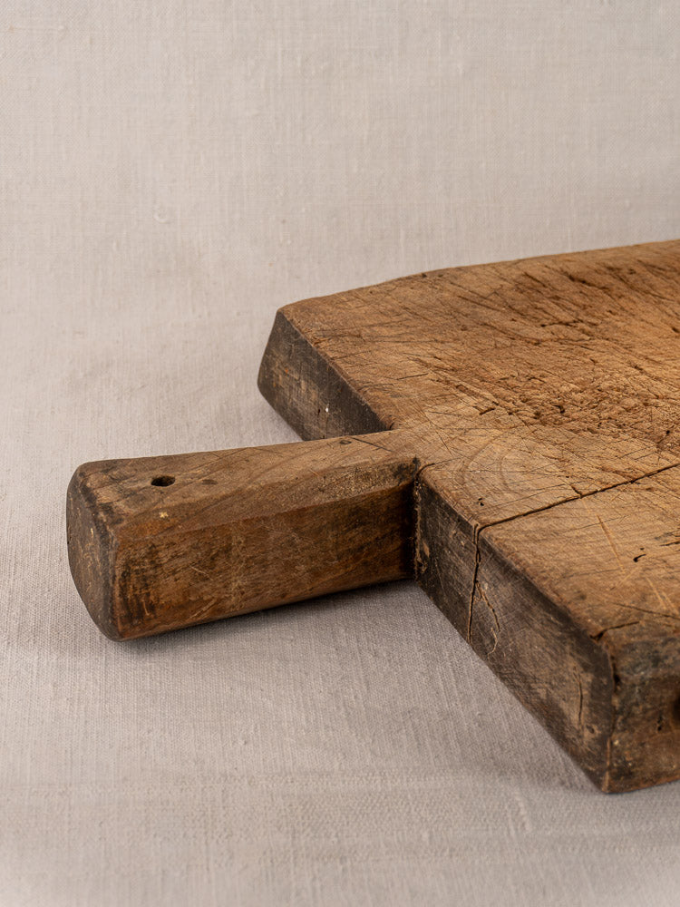 Antique French hand-carved cutting board with short handle, visible wear, on a light fabric background.