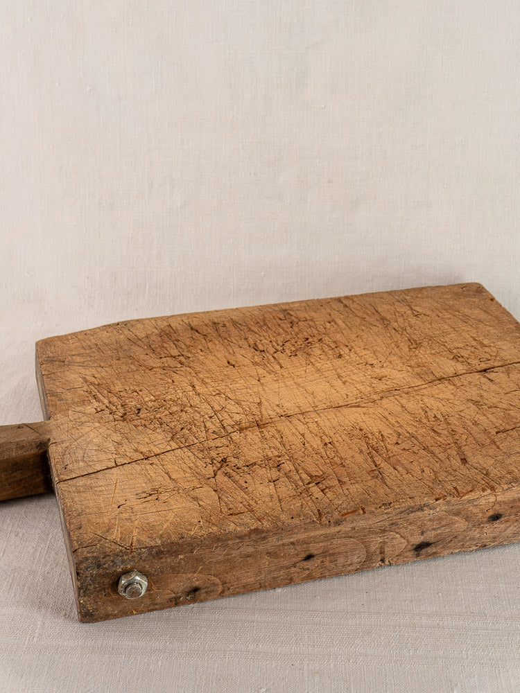 Antique French hand-carved cutting board with knife marks and metal bolt, displayed on neutral fabric background.