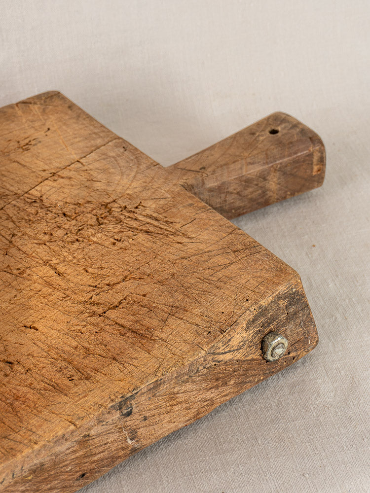 Antique French cutting board with deep knife marks, carved handle, metal bolt; displayed on light beige fabric.