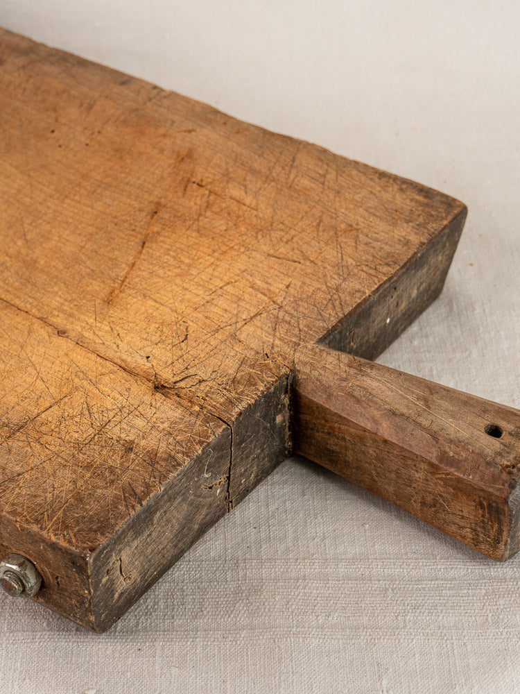 Antique French hand-carved cutting board with rectangular form, handle, deep scratches, and crack on aged surface over light fabric.