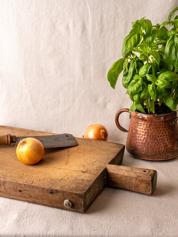 Antique French hand-carved cutting board with small knife, yellow onions, and copper pot with basil on neutral fabric.