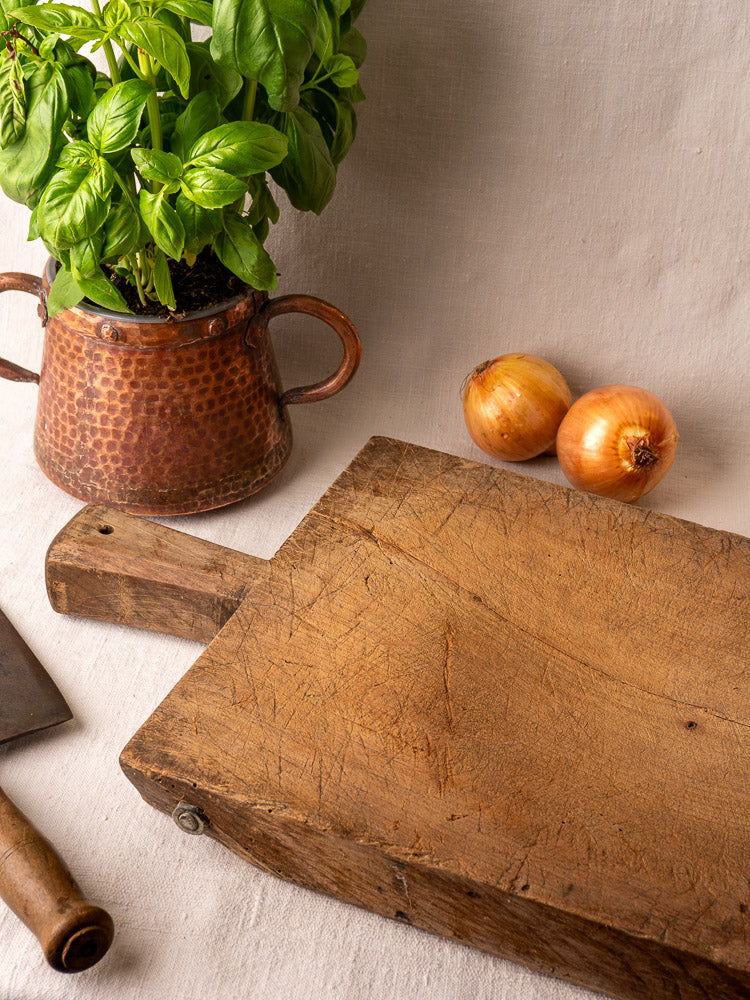 Antique French hand-carved cutting board on beige cloth with onions, metal-handled knife, and copper pot of basil.