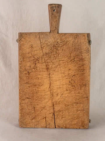 Antique French cutting board, hand-carved with handle, deep knife marks and crack, shown on neutral fabric.