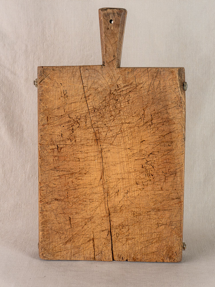 Antique French cutting board, hand-carved with handle, deep knife marks and crack, shown on neutral fabric.