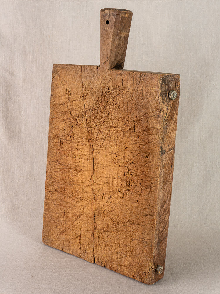 Early 19th-century antique French hand-carved cutting board with handle, upright against neutral fabric, visible wear and patina.