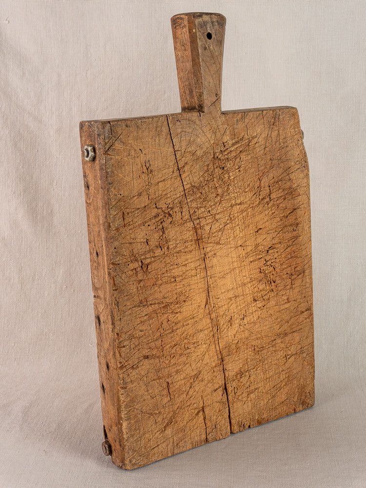 Antique French cutting board with handle, deep scratches, and large crack on neutral fabric background. Early 19th century.