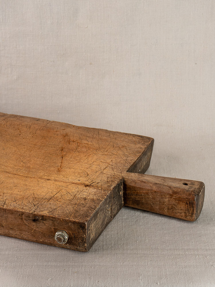 Antique French cutting board with thick rectangular shape, short handle, rich wood grain, on light fabric surface.