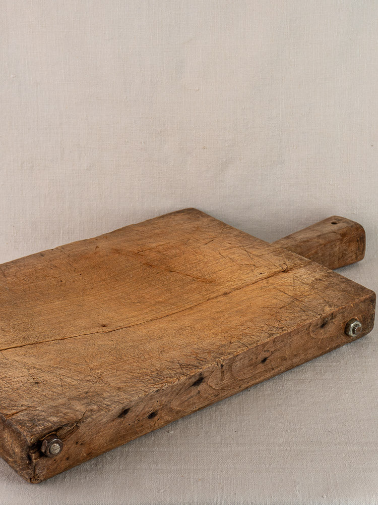 Antique French cutting board, hand carved with handle and metal bolts, resting on light beige fabric background.