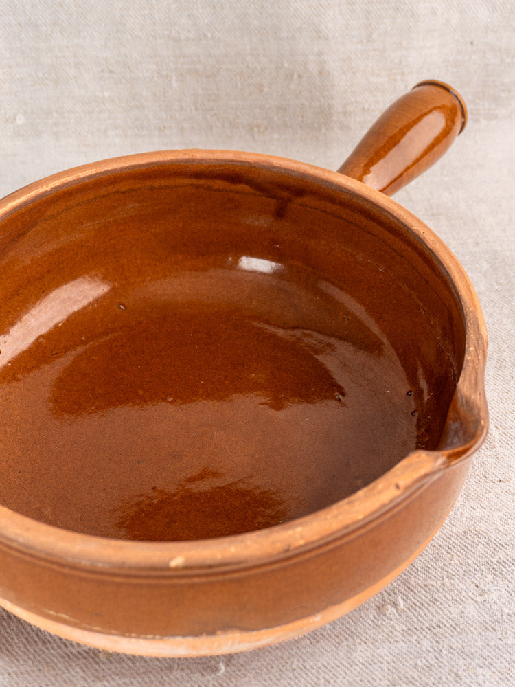 Antique French poêlon in glossy brown glazed terracotta with hollow handle and small spout on light fabric.