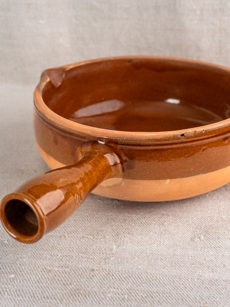 Mid-century French antique poêlon, round brown glazed terracotta pan with hollow handle, on beige textured fabric.