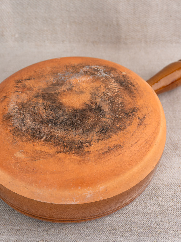 Antique French glazed terracotta poêlon with hollow handle, upside down on fabric, base shows black stains and burn marks.