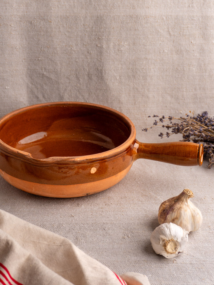 Antique French poêlon with glazed terracotta finish and hollow handle on beige cloth, beside garlic and dried lavender.