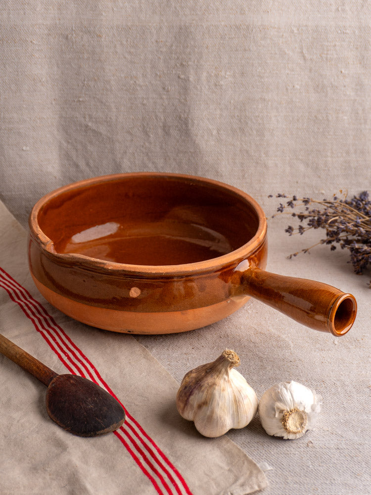Antique French brown glazed terracotta poêlon with hollow handle, on cloth beside garlic, wooden spoon, and striped napkin.