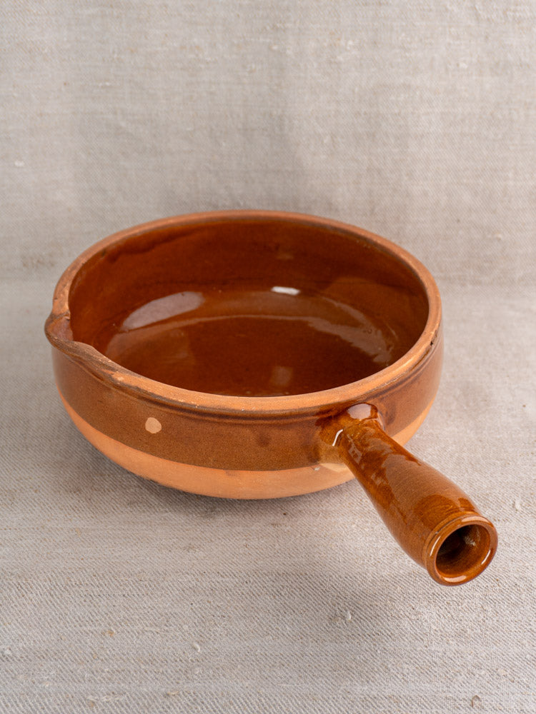 Mid-20th century French poêlon in glazed terracotta with glossy brown finish, hollow handle, and spout on beige fabric.