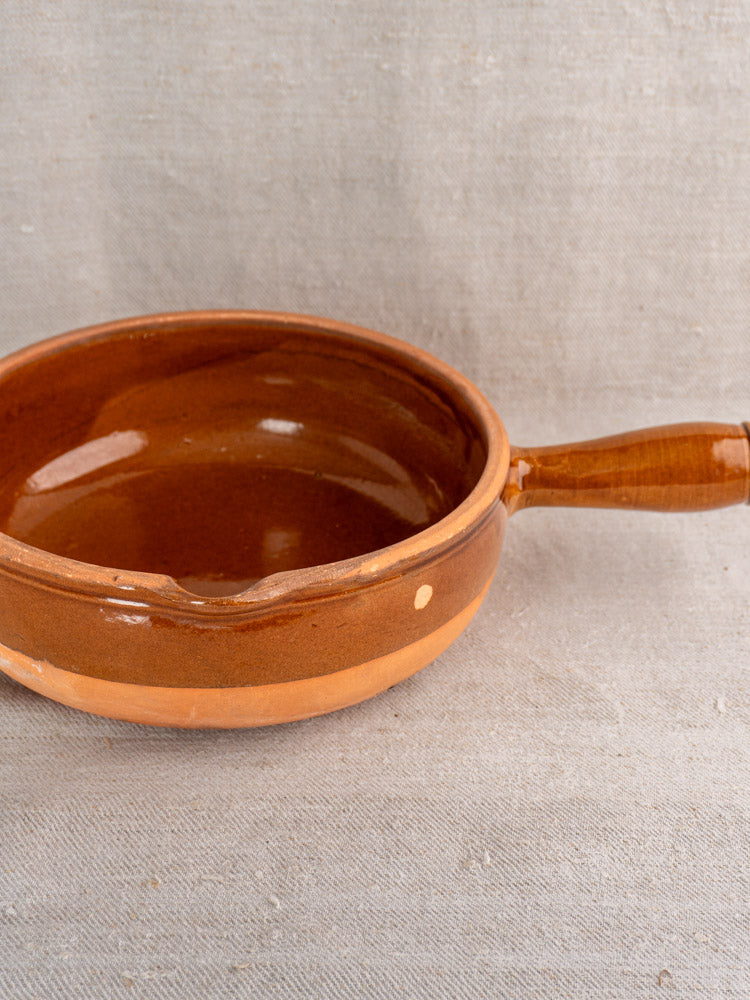 Mid-20th-century French glazed terracotta poêlon with hollow handle on light beige fabric surface, side view.