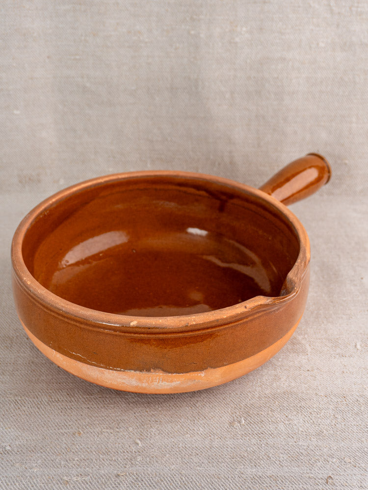 Mid-20th century French glazed terracotta poêlon with hollow handle, brown finish, on beige fabric background.