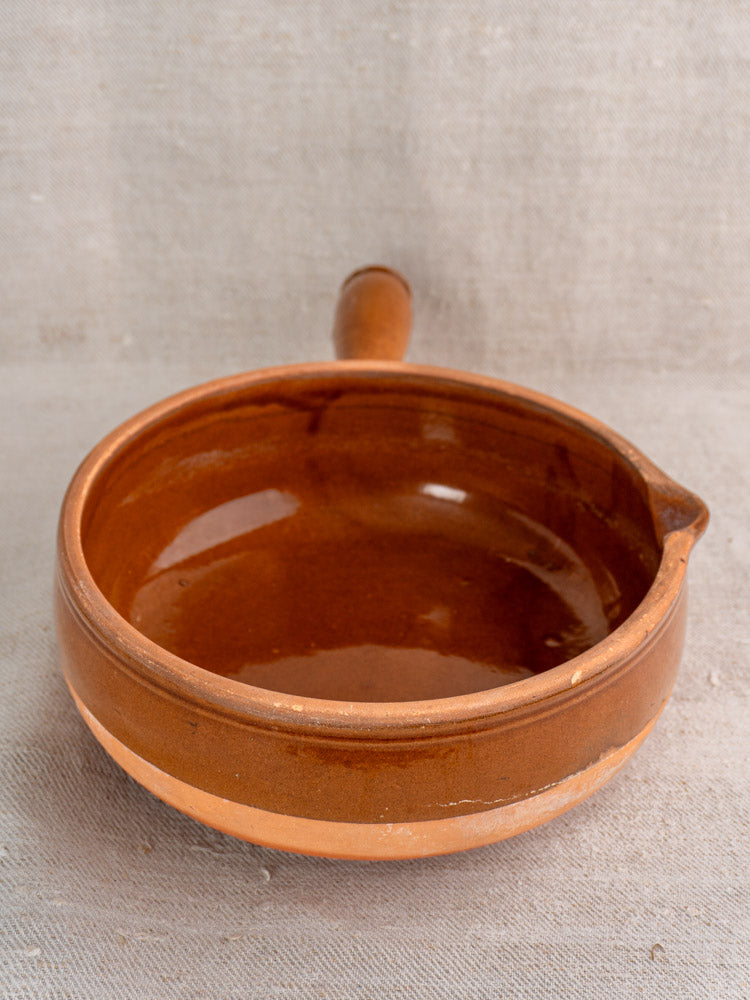Mid-20th-century French brown glazed terracotta poêlon with hollow handle and spout on textured light fabric.