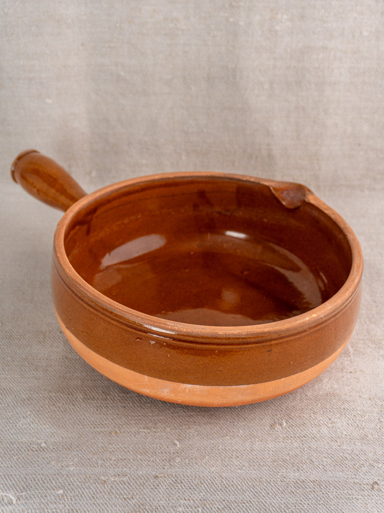 Antique French poêlon in glazed terracotta with hollow handle, mid 20th century, on beige fabric background.