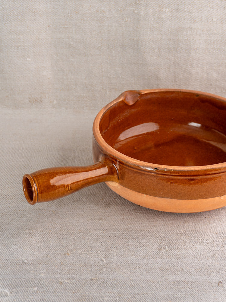 Mid-20th century French glazed terracotta poêlon with hollow handle on textured beige fabric.