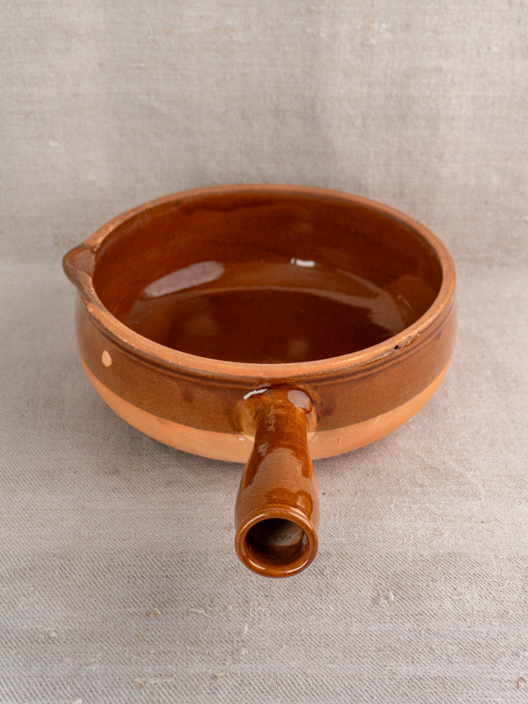 Mid-20th century French glazed terracotta poêlon pan with spout and hollow handle on beige fabric.
