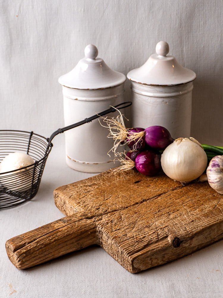 Antique French primitive cutting board with onions, garlic, and two white ceramic canisters nearby.