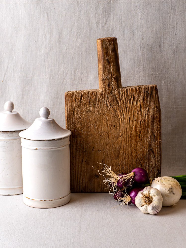 Antique French primitive cutting board with two white ceramic jars, garlic, and onions on fabric.