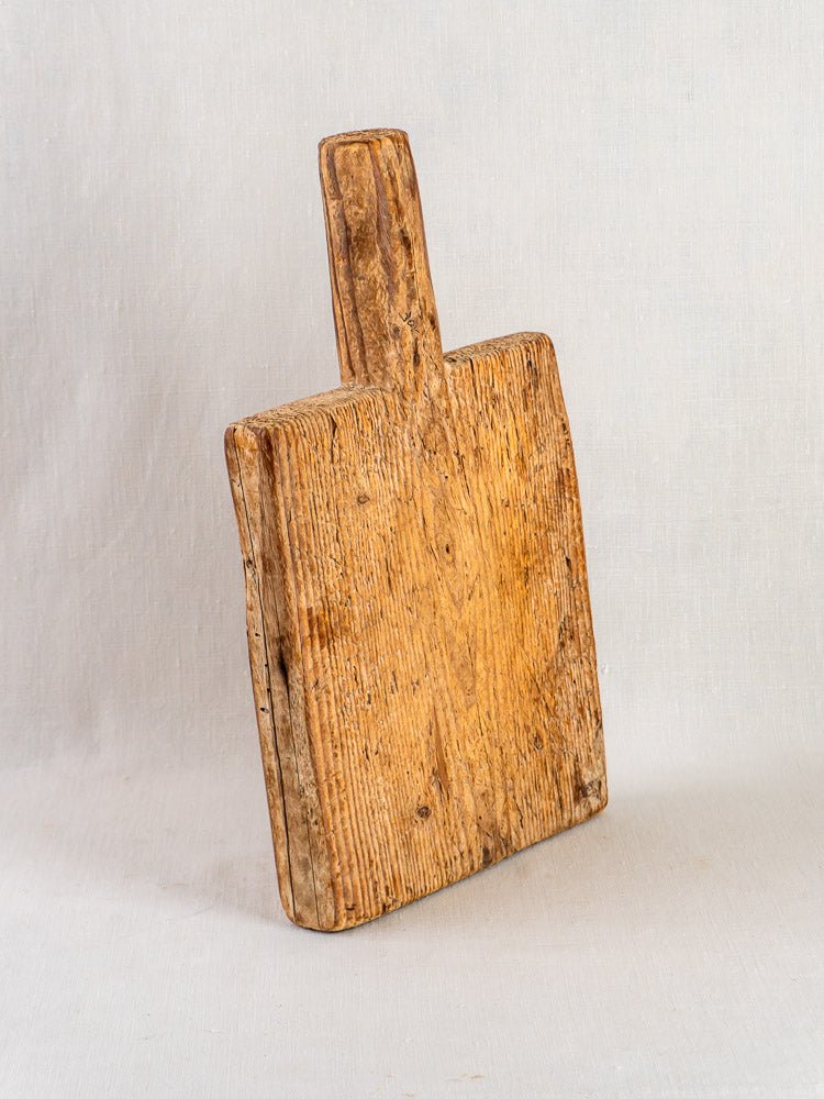 Antique 18th-century French primitive cutting board with handle, weathered texture, and visible wood grain on neutral fabric.
