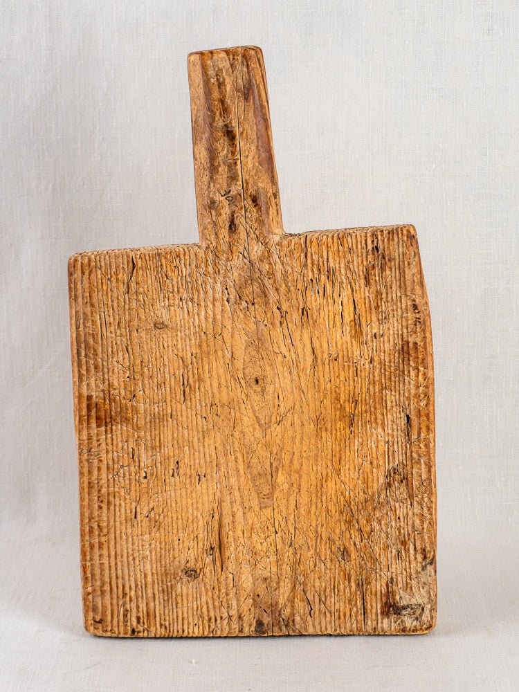 Antique 18th-century French primitive cutting board with short handle and vertical grain, light background.