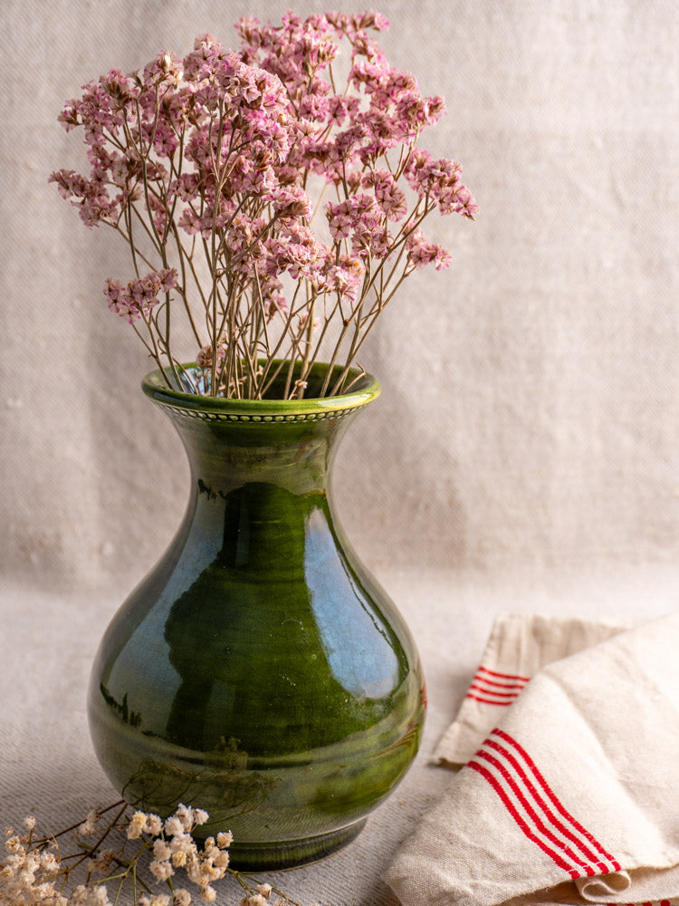Vintage French Pichon Uzès green glazed earthenware vase with dried pink flowers on beige fabric, red-striped cloth behind.