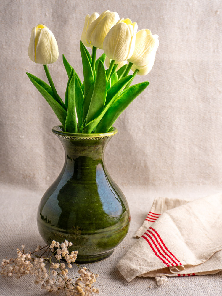 Vintage French Pichon Uzès green glazed earthenware vase with white tulips, on pale fabric beside striped cloth and dried flowers.