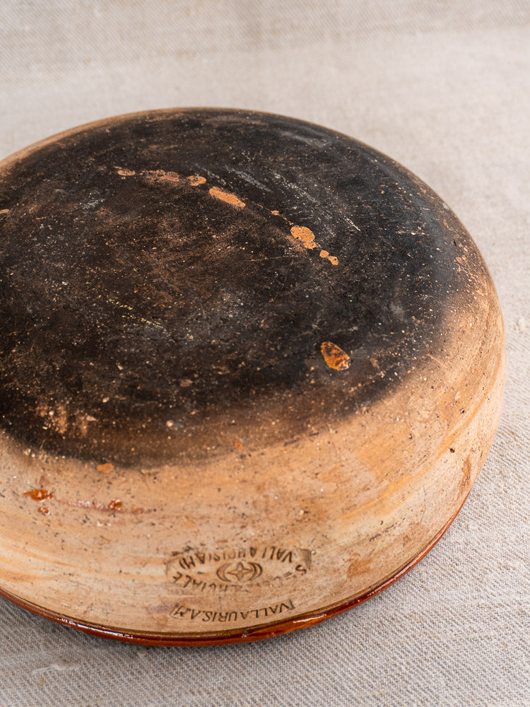 Underside of vintage French Vallauris terracotta poêlon with visible maker’s mark on pottery, fabric backdrop.