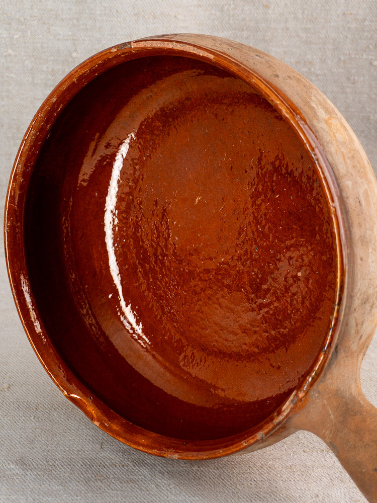 Vintage French Vallauris terracotta poêlon with handle, rustic brown glaze and shiny reddish interior on light gray textured surface.
