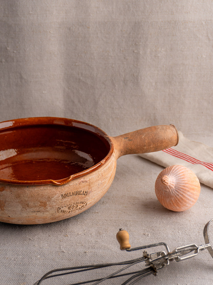 Vintage French Vallauris terracotta poêlon with handle, beside onion and hand mixer on beige fabric.
