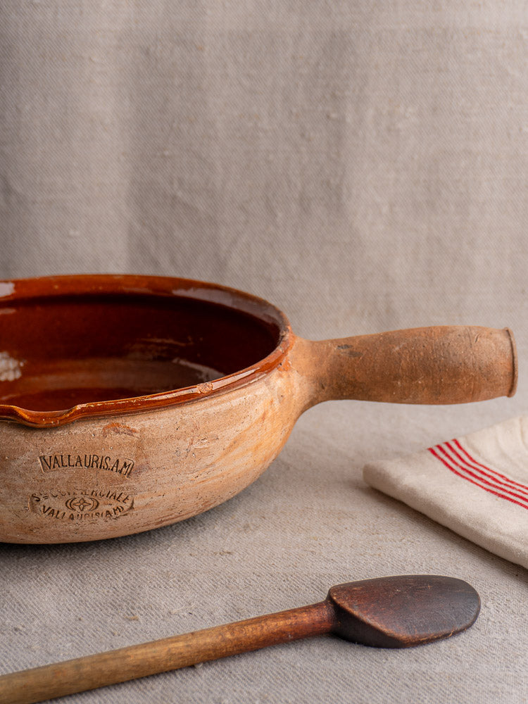 Vintage French Vallauris terracotta poêlon with sturdy handle beside red-striped cloth and wooden spoon.