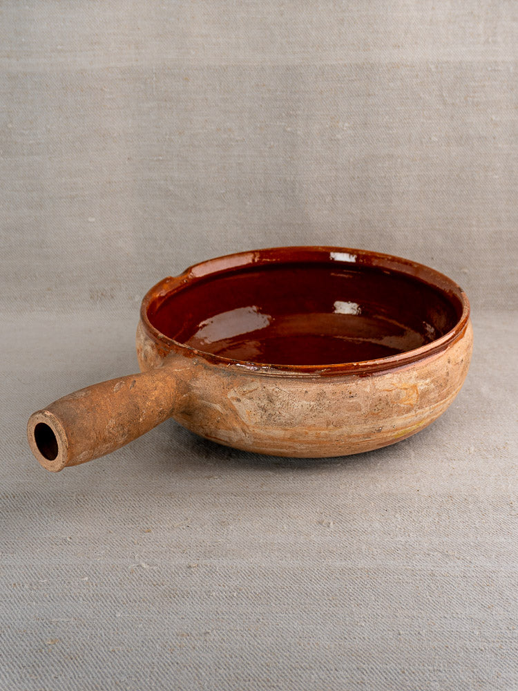 Vintage French Vallauris terracotta poêlon with short handle and reddish-brown glazed interior on neutral fabric.
