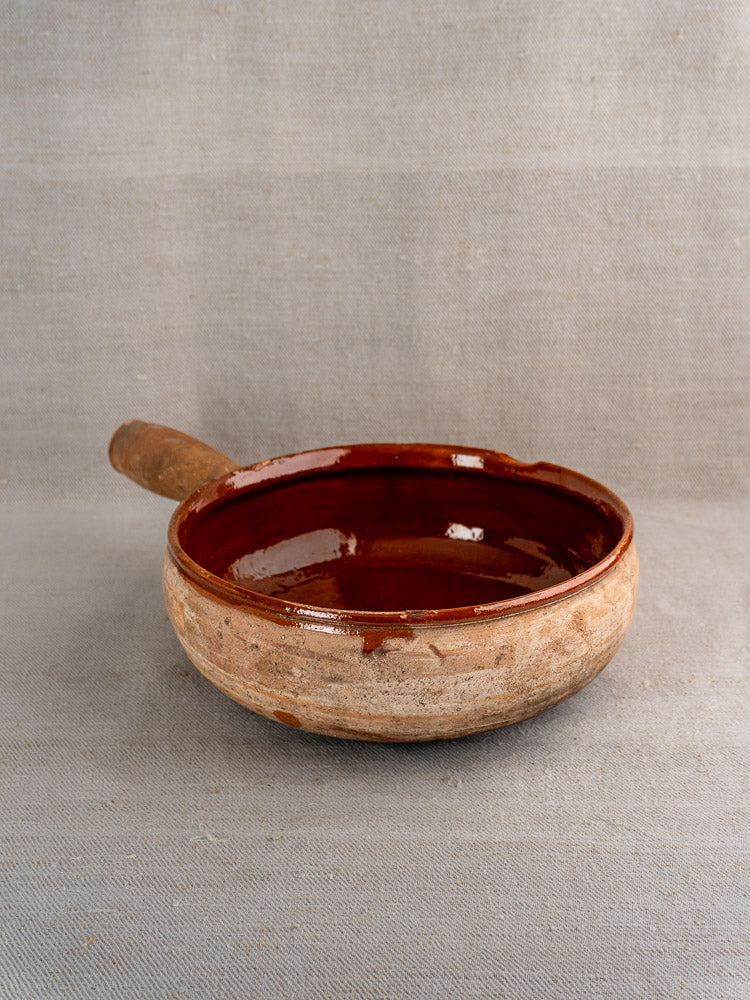 Vintage French Vallauris terracotta pan with handle, dark glazed interior, unglazed beige exterior on gray surface.