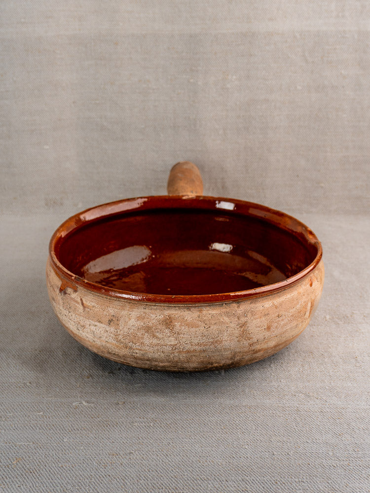 Vintage French Vallauris poêlon, round terracotta pan with glossy brown glaze, on gray fabric.