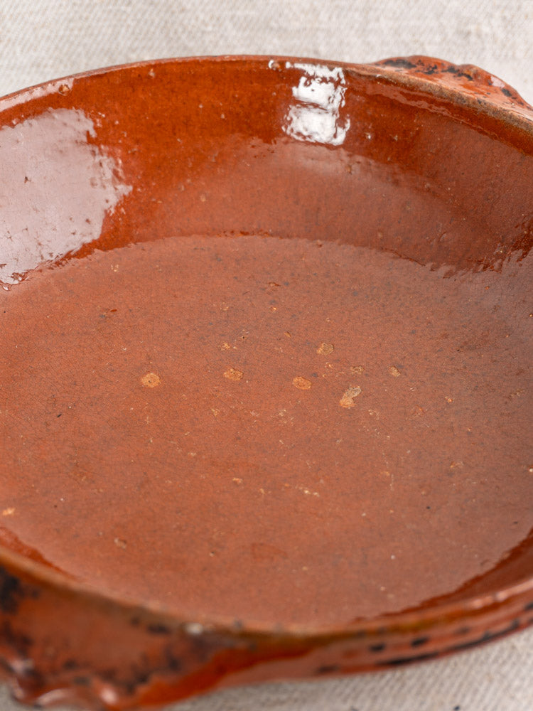 Rustic Mid-Century Vallauris terracotta baking dish with handles, round shallow form, glazed, on light fabric background.