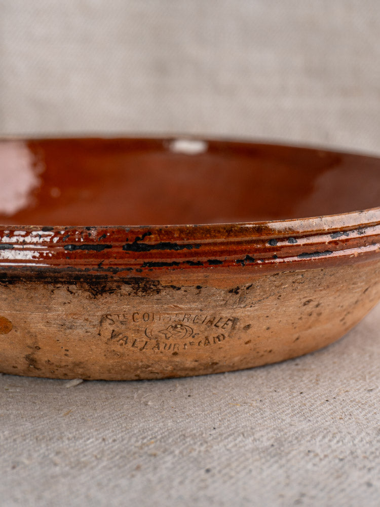 Mid-century Vallauris terracotta baking dish with handles, reddish-brown glazed interior, maker’s mark, on neutral woven fabric.