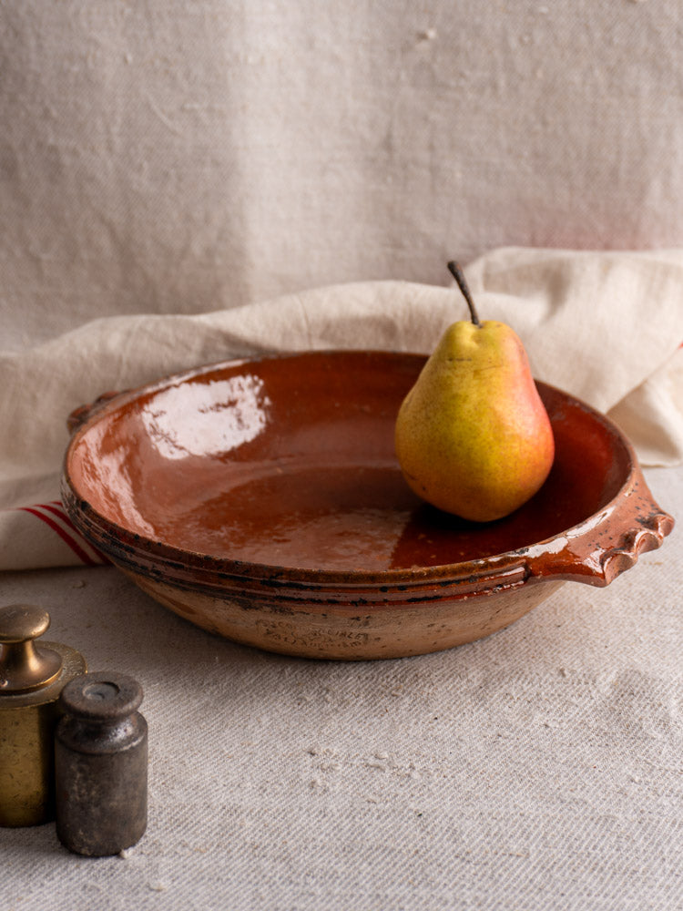 Rustic Mid-Century Vallauris terracotta baking dish with handles, displayed with brass weights on textured fabric.