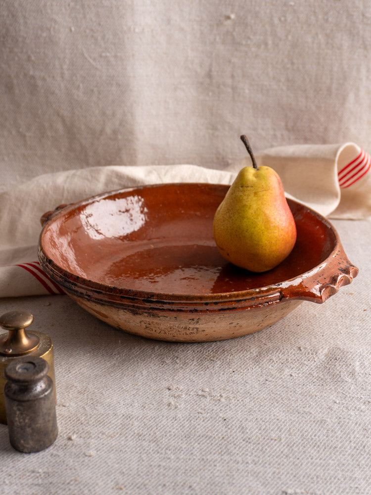 Mid-century Vallauris terracotta baking dish with handles, shown on textured beige fabric with pear and red-striped cloth.